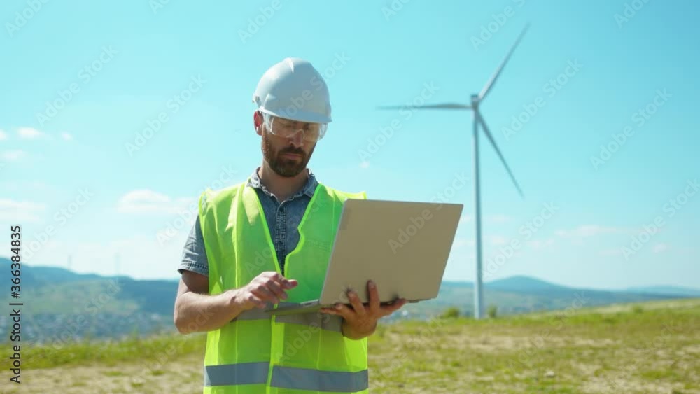 In uniform man engineers use laptop stand near windmills in fields labor collaboration contribution windpower farm technicians close up slow motion In uniform man engineers use laptop stand near windmills in fields labor collaboration contribution windpower farm technicians close up slow motion