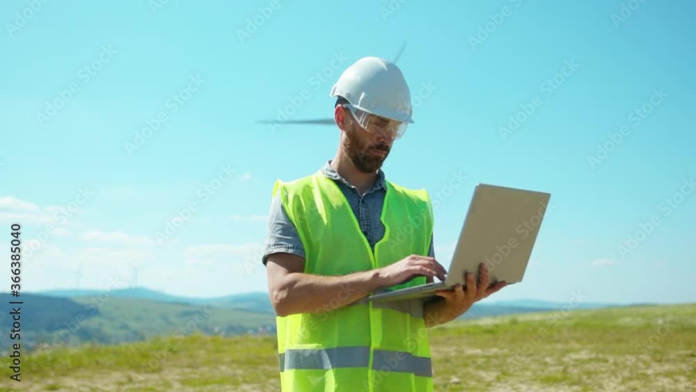 Portrait young man engineers in uniform use laptop stand near windmills in fields labor collaboration contribution windpower farm technicians close up slow motion Portrait young man engineers in uniform use laptop stand near windmills in fields labor collaboration contribution windpower farm technicians close up slow motion