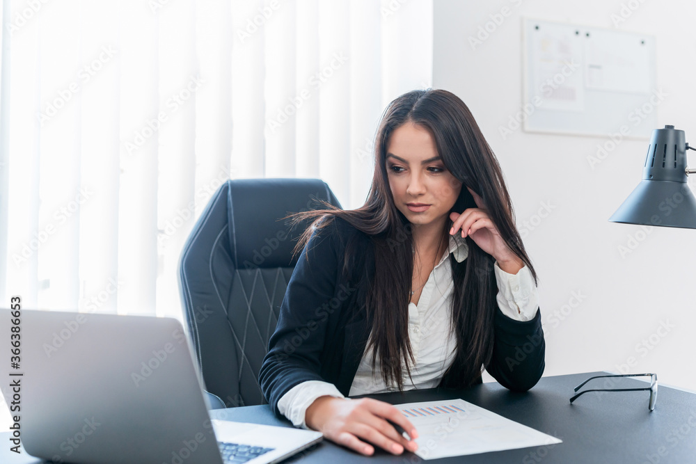 Business woman with long brown hair sitting at desk and looking at laptop. Business woman with long brown hair sitting at desk and looking at laptop.