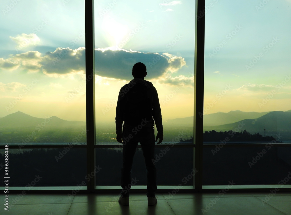 Fototapeta premium Hombre mirando el atardecer en un ventanal con la ciudad montañas de fondo