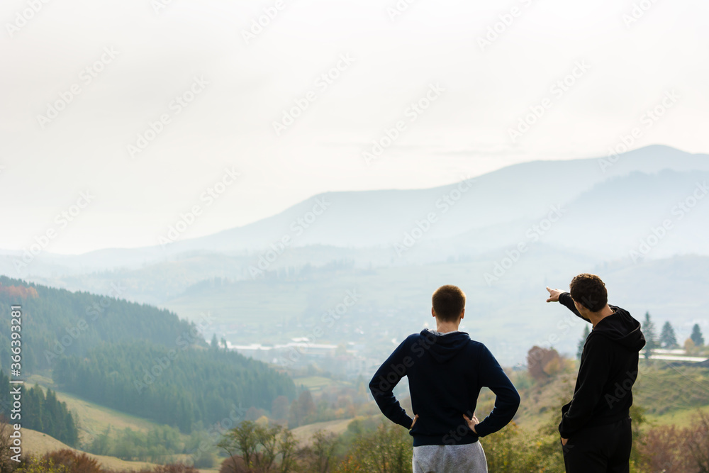 Naklejka premium The guys stand looking at the mountains covered with fog and plan their ascent. Summer. Camping. Picnic. Tourism. Green colors. Daylight. A city in the distance.