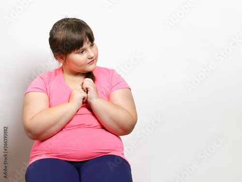 Little plump girl sits and looks to the side in thought. Problem of excess weight in children, after quarantine. On light background with copy space