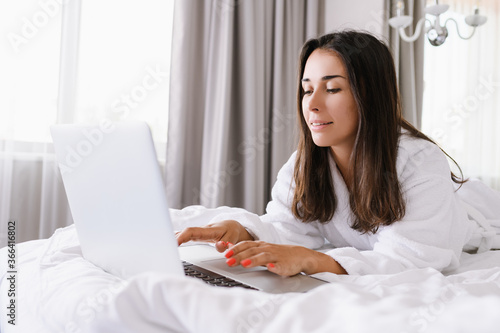 Beautiful girl lay in white bed in morning light, wear bathrobe, chatting, network online, type at laptop keyboard, freelancer, business, shopping online, social net concept © o_lypa