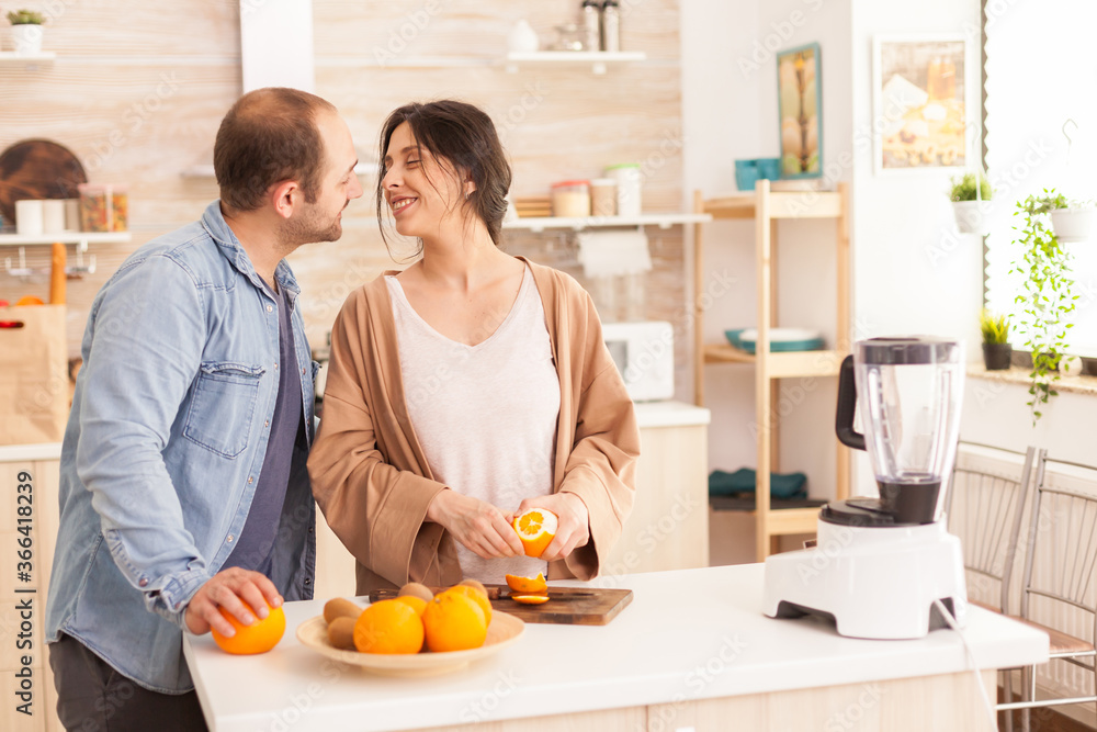 Couple making smoothie in kitchen . Woman peeling of orange while smiling at husband. Healthy carefree and cheerful lifestyle, eating diet and preparing breakfast in cozy sunny morning Couple making smoothie in kitchen . Woman peeling of orange while smiling at husband. Healthy carefree and cheerful lifestyle, eating diet and preparing breakfast in cozy sunny morning