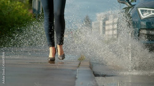 SLOW MOTION, LOW ANGLE, CLOSE UP, DOF: Unrecognizable young woman wearing high heels gets splashed with water as inconsiderate driver drives their car into a deep puddle at the side of the road. photo