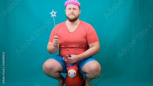 handsome bearded freaky man in a pink T-shirt with a deadema on his head is saddened riding a unicorn with a magic wand in his hand. A funny wizard joke to make and fulfill a wish. photo