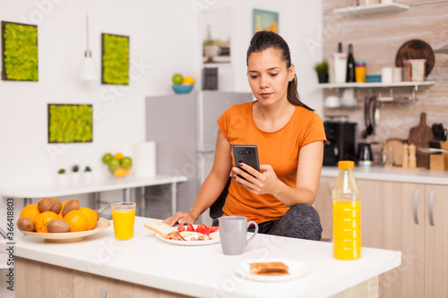 Woman checking social media in the morning at breakfast time. Housewife using modern tehcnology and drinking healthy, natural, homemade orange juice. Refreshing morning.