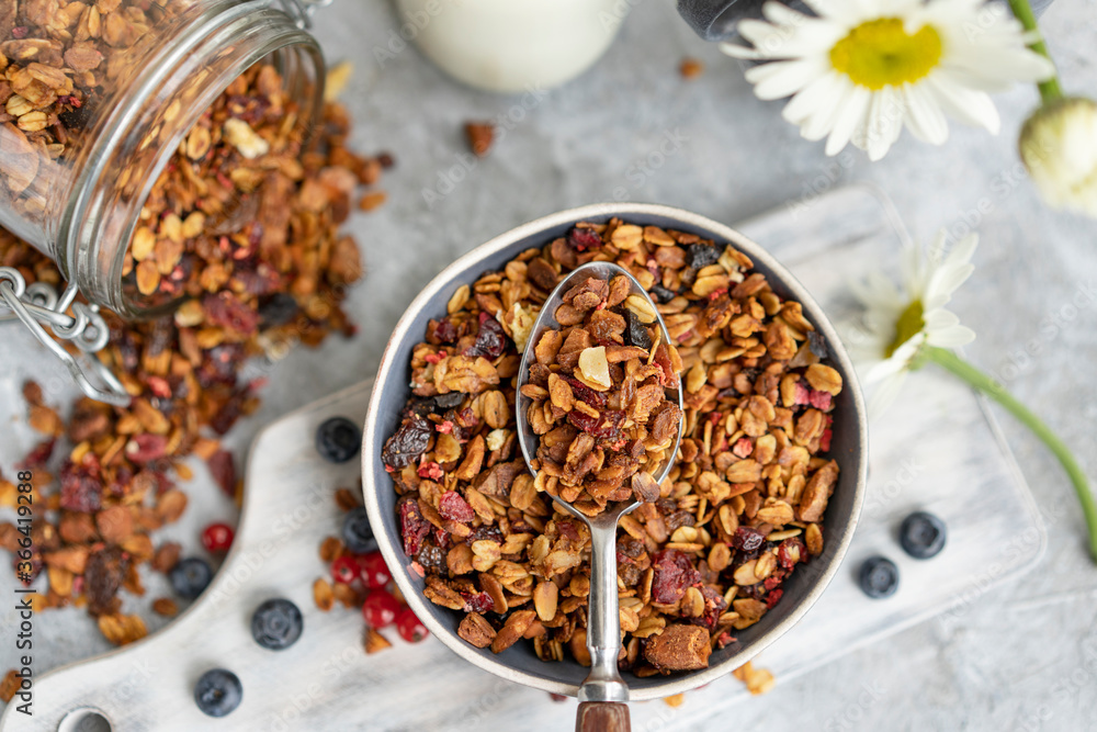 Fototapeta premium Homemade granola or muesli made of oats, nuts, honey, cinnamon, fruits on white table with milk and flowers. Beautiful healthy snack on breakfast. Top view, flat lay, copy space.