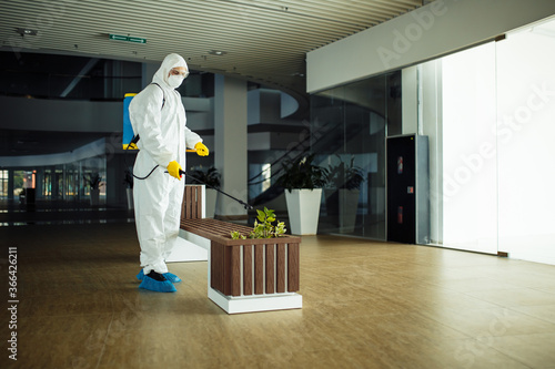 A man wearing protective suit is disinfecting a bench in an empty shopping mall with sanitizing spray. Cleaning up the public place to prevent covid spread. Healthcare precautions and safety concept.