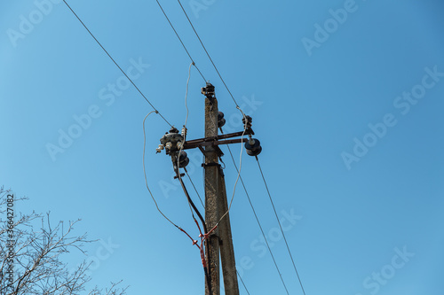  Power line post with electricity cables against a clear sky with white clouds. A tree grows near a pillar. Electricity transmission line, power supply