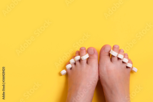 Female feet with sponge for pedicure on fingers on bright yellow background with copy space