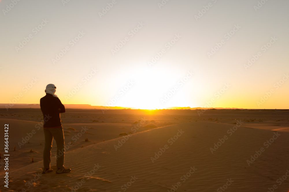 A man alone in a turban with his arms crossed watches the sunrise in the Erg Chebbi desert in Merzouga, Morocco