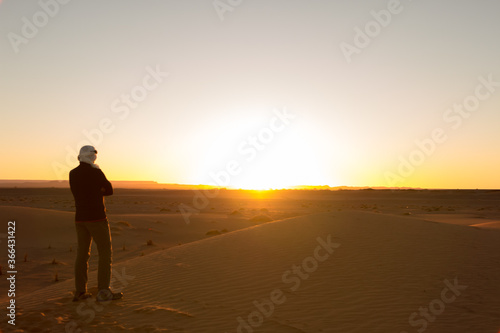 A man alone in a turban with his arms crossed watches the sunrise in the Erg Chebbi desert in Merzouga  Morocco