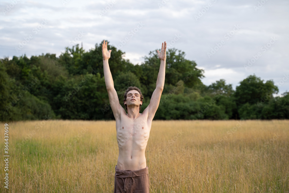 Shirtless man with eyes closed and arms raised exercising on landscape against sky in forest Shirtless man with eyes closed and arms raised exercising on landscape against sky in forest