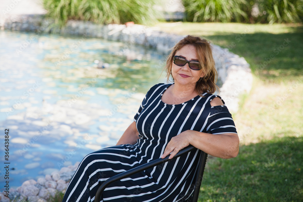 Portrait of lovely middle aged woman with sunglasses sitting on the chair at the park. Image of relaxed senior woman sitting outdoor.