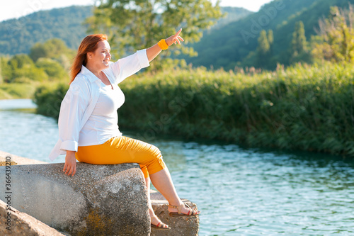 A happy overweight woman looks into the distance and points her finger while sitting on a sea pier. Copy space. Side view