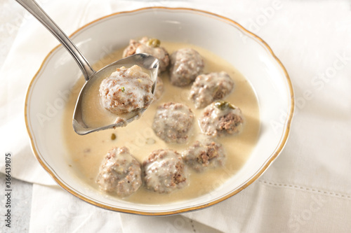 Ladle is liftiting a meatball from a bowl with koenigsberger klops, which are boiled beef balls in a white bechamel sauce with capers, traditional Polish and German dish, selected focus photo