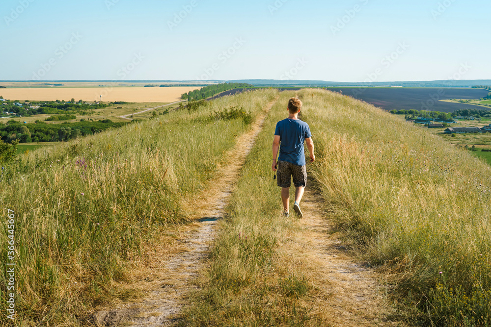 A young male tourist walks along an overgrown path in the mountains overlooking fields