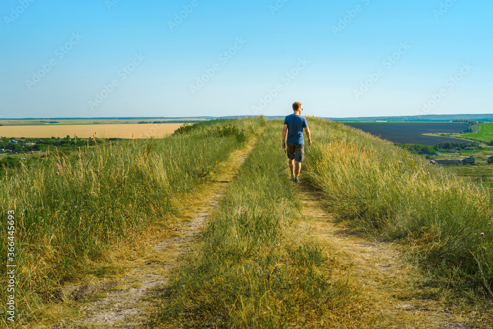 A young male tourist walks along an overgrown path in the mountains overlooking fields