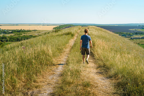 A young male tourist walks along an overgrown path in the mountains overlooking fields