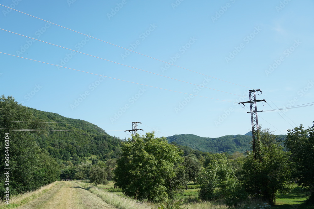 A footpath among rich vegetation. Region of Zarnovica in central Slovakia. The lanscape is crossed by electric wires and by masts or pillars for transmission of electricity.