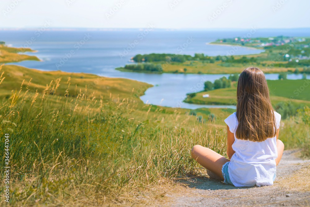 Little girl child with long hair sitting on a path with a mountain landscape with rivers and green meadows, photo from the back