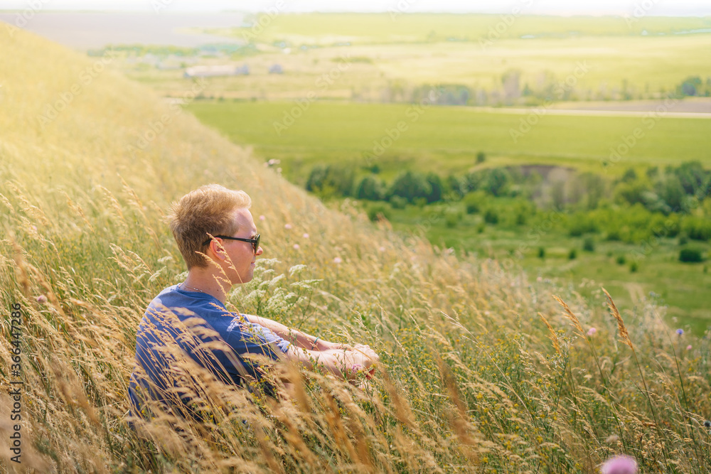 A young male tourist camped in the mountains, sitting on the grass with a view of the mountains and meadows