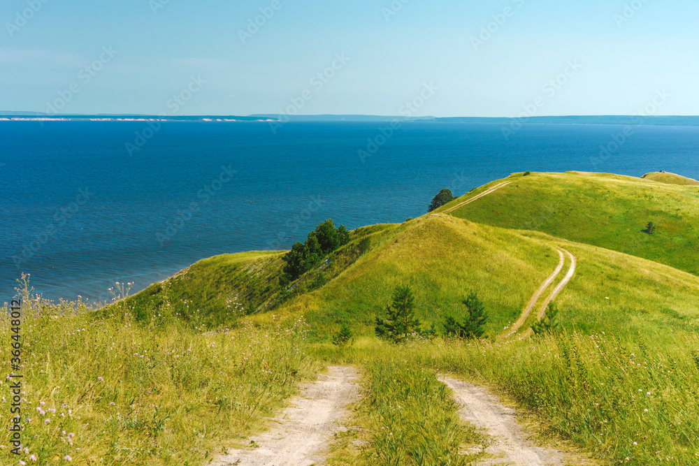 A wide shot of a curved gravel road through a lush green hillside with the sea or lake in the background.