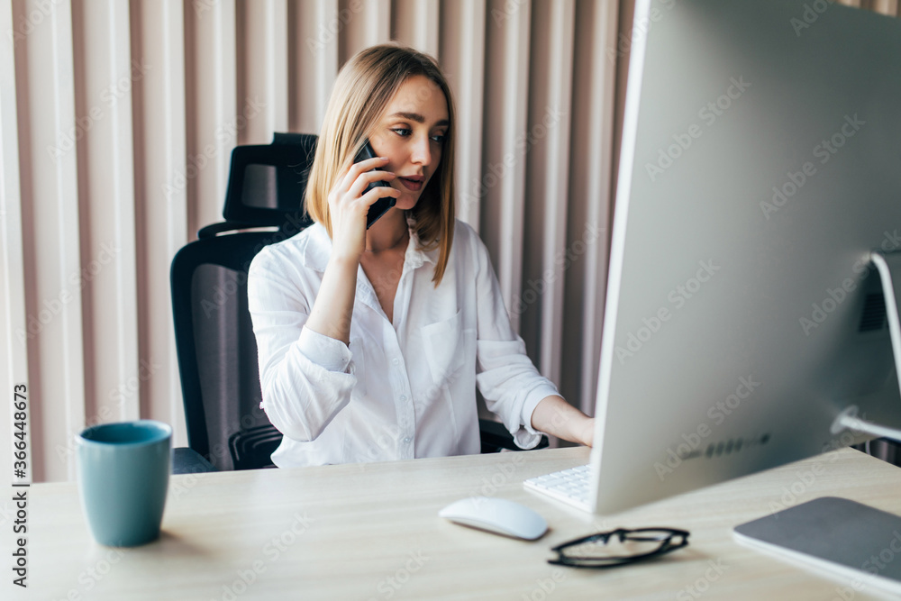 Portrait of beautiful happy smiling young designer woman working on laptop at office desk. Attractive cheerful model in suit using smartphone and computer, making call and typing. Indoors Portrait of beautiful happy smiling young designer woman working on laptop at office desk. Attractive cheerful model in suit using smartphone and computer, making call and typing. Indoors