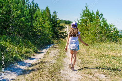 A happy little girl a child with a bandana and denim shorts is walking along a path dancing through green mountain meadows, hiking