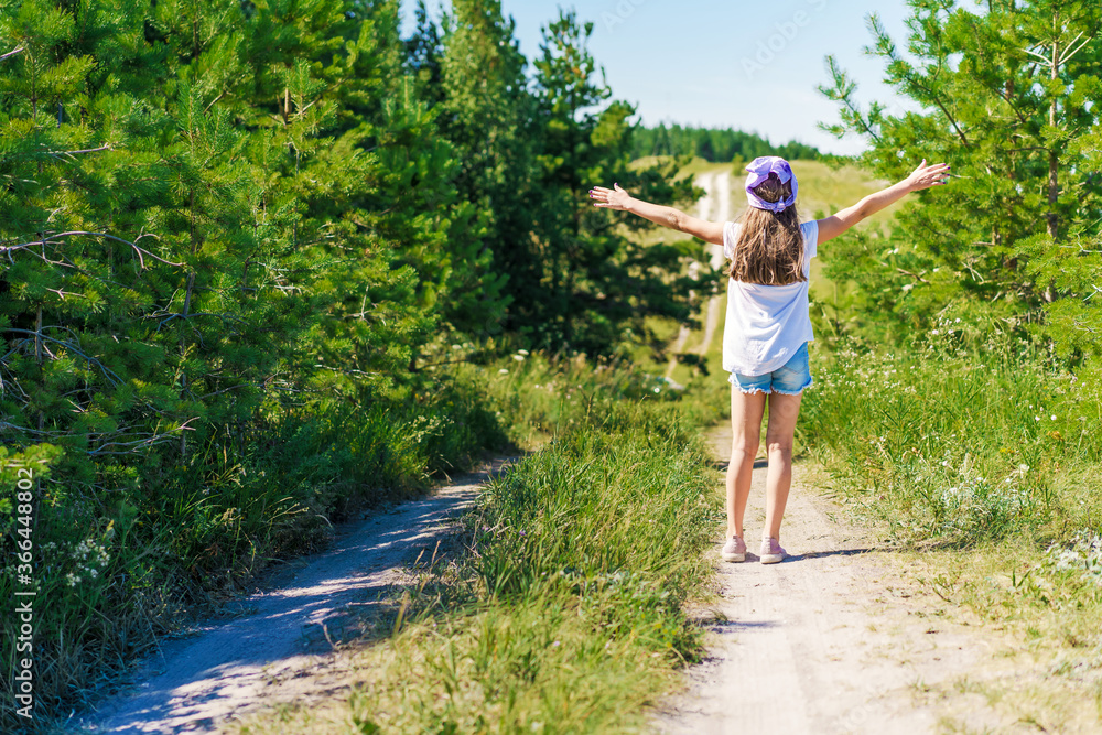 A happy little girl a child with a bandana and denim shorts is walking along a path dancing through green mountain meadows, hiking