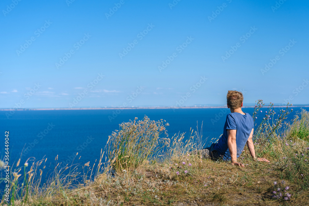 A blond man in a blue shirt sits on a cliff overlooking the blue sea and Islands