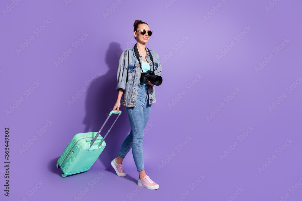 Full length profile side photo of positive cheerful girl travel weekend tourism go walk step departure hold suitcase wear denim jeans jacket singlet isolated violet color background Full length profile side photo of positive cheerful girl travel weekend tourism go walk step departure hold suitcase wear denim jeans jacket singlet isolated violet color background