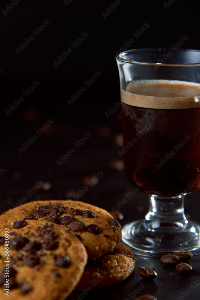 Close up of a glass cup of coffee, chocolate chip cookies and roasted coffee beans on dark background. Concept of ready to eat food, tasty snack. Selective focus, copy space. Close up of a glass cup of coffee, chocolate chip cookies and roasted coffee beans on dark background. Concept of ready to eat food, tasty snack. Selective focus, copy space.