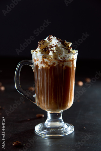 Close up of a glass cup of coffee with whipped cream and shaved chocolate on it and roasted coffee beans on dark background. Concept of ready to eat food, tasty snack. Selective focus, copy space.
