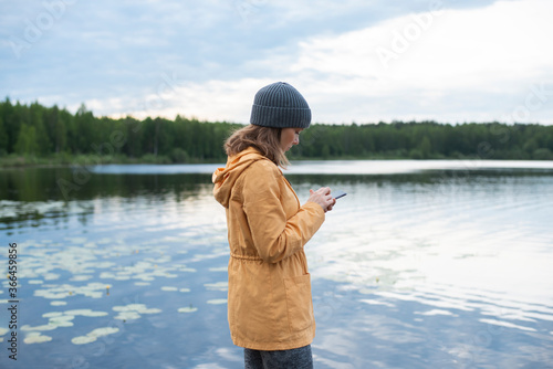 Young woman using smartphone while standing at lakeshore against cloudy sky
