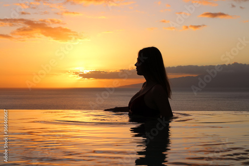 A girl in a swimsuit with her hair down stands sideways and looks at the sunset.