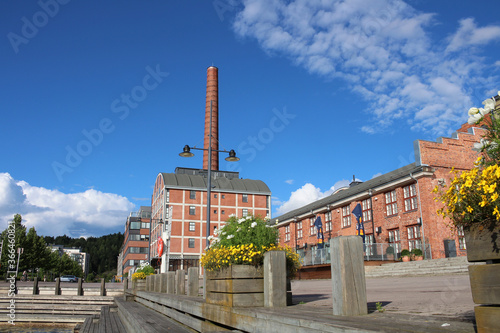 Wooden embankment with steps and flower beds against the background of a red brick factory with a large chimney on the shore of lake Vesiyarvi in the city of Lahti. Finland.