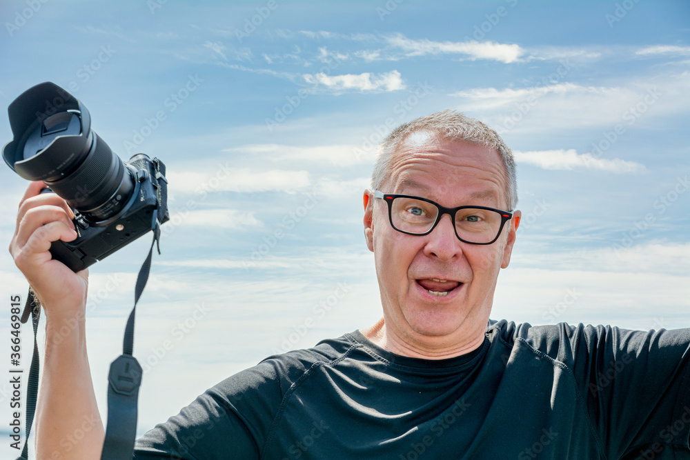 Obraz premium A middle-aged man, in his 50th, holds up a camera, looks straight into the camera, and looks happy. Blue hazy sky background.