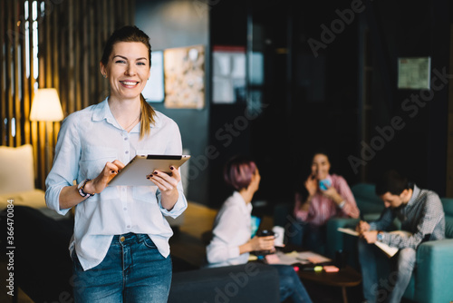 Half length portrait of cheerful young woman in casual wear standing with portable pc for work in office,prosperous female leader of professional satisfied with successful career looking at camera.