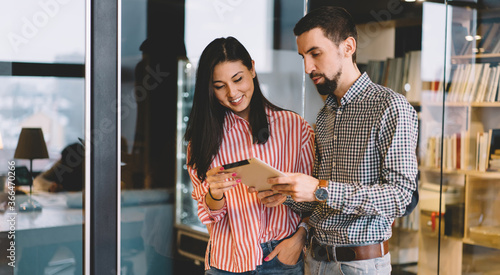 Bearded male employee showing his female colleague new modern application for digital devices on tablet, positive asian woman watching video on portable pc and communicating with friend on break