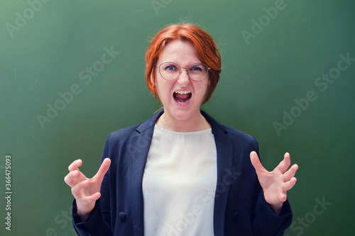 Woman teacher screams furious at the blackboard, copy space background. Angry school teacher on green background, close up