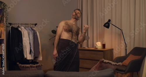 Good looking mature guy in bath towel pretending playing guitar while brushing teeth. Emotional bearded man in 40s having fun and singing at home. Concept of happy people. photo