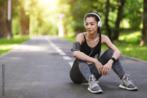 Rest After Training Outdoors. Young Asian Woman In Headphones Sitting On Path