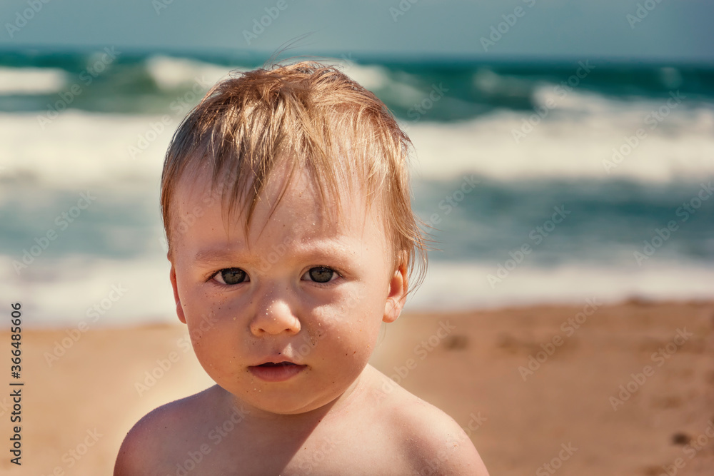 Adorable infant boy looking at camera at summertime. Young caucasian baby at tropical beach with ocean wave behind him. Male kid portrait on his first vacations at sandy beach. Curiosity expression. Adorable infant boy looking at camera at summertime. Young caucasian baby at tropical beach with ocean wave behind him. Male kid portrait on his first vacations at sandy beach. Curiosity expression.
