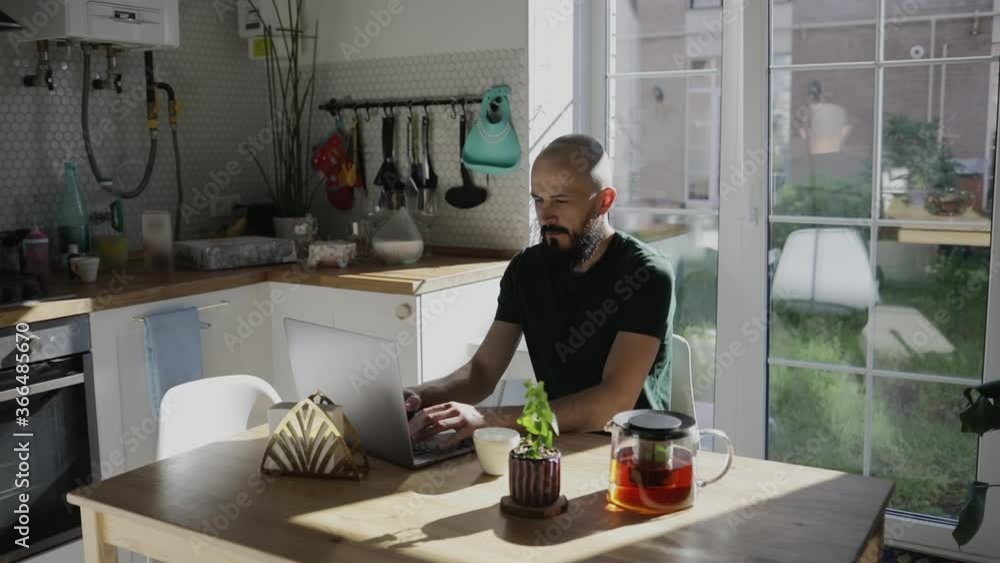 Freelancer programmer working from home on kitchen and using laptop. Bearded man working with a computer, prints text on keyboard. Self entrepreneur sitting and working at his modern apartment. Freelancer programmer working from home on kitchen and using laptop. Bearded man working with a computer, prints text on keyboard. Self entrepreneur sitting and working at his modern apartment.