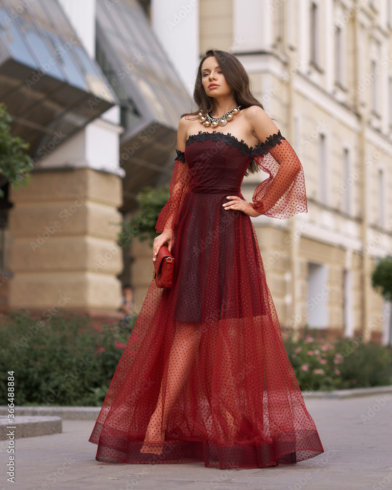 Elegant fashionable female model in red long evening dress with open shoulders and sleeves standing and posing at city street. Beautiful caucaisan woman with wavy hair and makeup outdoors portrait