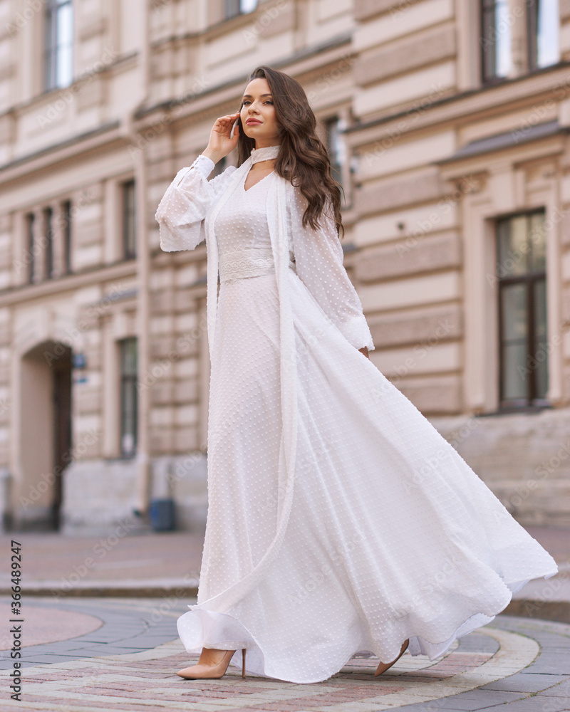 Elegant fashionable stylish female model in long white dress walking at city street. Outdoor full length portrait. Caucasian lady with wavuy hair and makeup