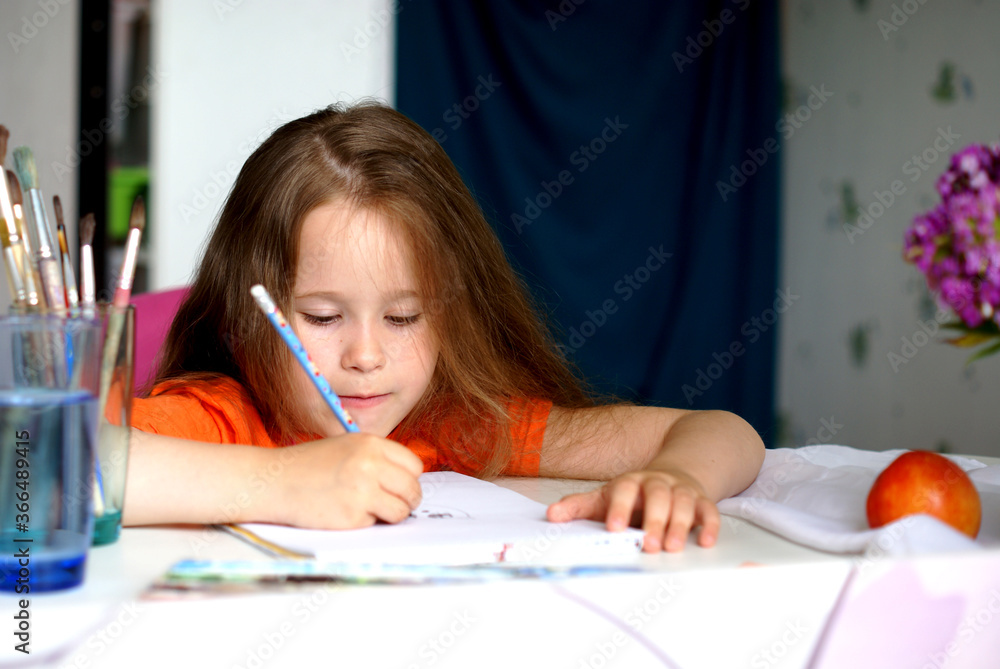 Caucasian child girl sitting at the table and draws with a pencil. Before her are paints and brushes Caucasian child girl sitting at the table and draws with a pencil. Before her are paints and brushes