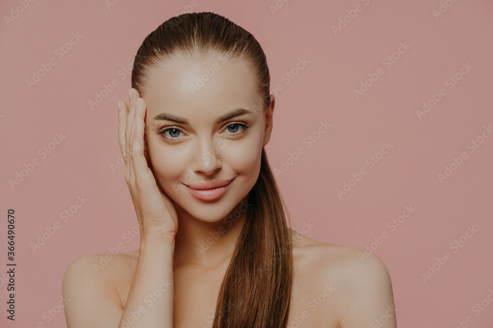 Isolated shot of pretty woman with combed pony tail, enjoys freshness of skin, looks with charming expression, stands bare shoulders, isolated on pink wall. Health care, makeup, spa and face lifting Isolated shot of pretty woman with combed pony tail, enjoys freshness of skin, looks with charming expression, stands bare shoulders, isolated on pink wall. Health care, makeup, spa and face lifting